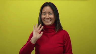 Kırmızı bir kazakla parlak sarı bir arka plana doğru mutlu bir şekilde el sallayan bir kadın izole edilmiş neşeli bir ortamda gençliğini ve pozitifliğini sergiliyor..