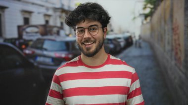 Sakallı ve gözlüklü genç adam arka planda park etmiş arabaları ve binaları olan bir sokakta gülümsüyor..