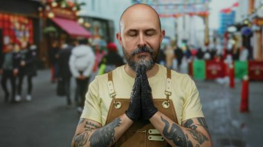 Dövmeli, kolları ve sakalı olan kel bir adam, şehir sokaklarında elleri sımsıkı bağlı, kahverengi önlük ve siyah eldivenlerle meditasyon yapıyor..