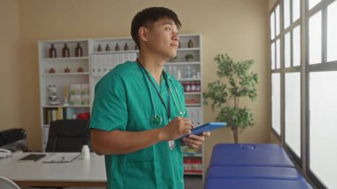 Çağdaş hastane kliniğinde tablet kullanan yeşil önlüklü genç adam profesyonel sağlık hizmetlerinin kapalı alanda olduğunu gösteriyor..