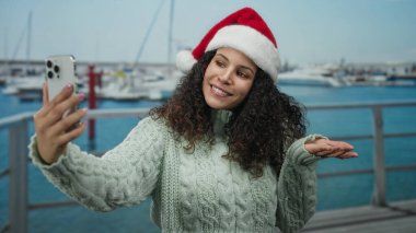 Noel Baba şapkalı bir kadın tatil sezonunda teknelerle deniz kenarında selfie çekiyor. Canlı bir açık hava manzarası ve şenlikli bir ruh hali sergiliyor..