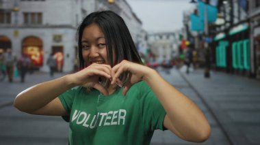 Yeşil gönüllü tişörtlü bir kadın şehir sokaklarında el ele gezerken kalp işareti oluşturuyor. Açık havada gençlik enerjisi ve toplum etkinliği..