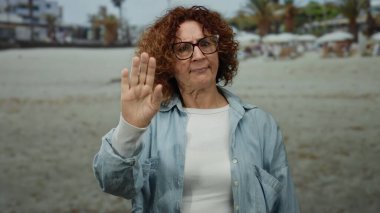 Sahilde el kol hareketleri yapan bir kadın, gözlük ve kot ceket takarak deniz kenarında bulutlu gökyüzünün altında, palmiye ağaçlarının yanında sıradan bir açık hava manzarası izlenimi uyandırıyor..