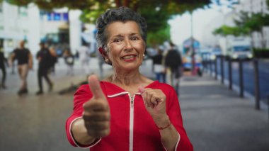 Kırmızı kazaklı, gri saçlı yaşlı bir kadın işlek bir şehir caddesinde başparmak kaldırıyor, bir şehir ortamında pozitifliği ve mutluluğu somutlaştırıyor.