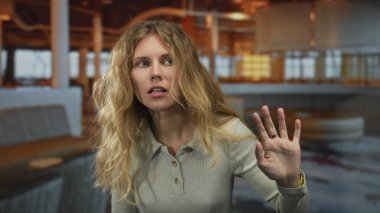 Kadın elini kaldırıyor, sallanıyor, sarışın genç bir figür gibi modern gündelik bir ortamda şaşırmış bir ifadeyle sesleniyor..