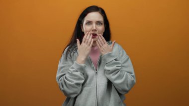Ağzı açık şaşırmış bir kadın gri kapüşonlu giyer parlak turuncu arka planda durur şok ve duygu taşır..