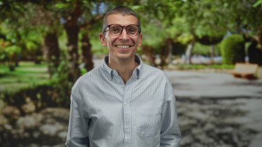 Güneşli bir parkta gülümseyen, gözlüklü ve çizgili tişörtlü, yemyeşil ağaçlar ve patikalarla çevrili, neşeli bir açık hava yaratan bir adam..