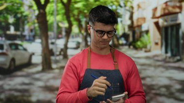 Kırmızı kazak ve kot önlük giyen İspanyol genç adam ağaçlarla ve park halindeki arabalarla dolu bir sokakta kalemle notlar alıyor.