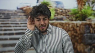 Sakallı ve çizgili gömlekli genç adam güneşli bir günde dışarıda taş basamaklar ve yeşilliklerle çevrili bir şekilde jest yapıyor..