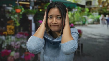 Woman standing outside a flower shop with hands on ears looking thoughtful surrounded by vibrant flowers and greenery capturing the essence of tranquility in urban outdoor setting.