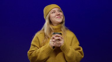 Elinde kahve fincanı tutan, sarı kazak ve şapkalı, mavi arka planda gülümseyen bir kadın..