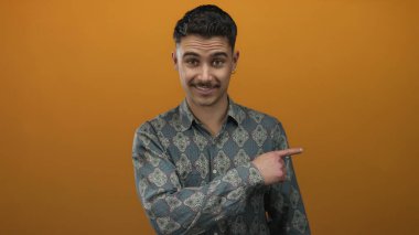 Şablonlu tişört giyen genç İspanyol adam canlı turuncu bir arka plana şaşırmış görünüyor, izole bir ortamda merak ve duygu yayıyor..