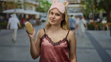 Gece elbiseli genç bir kadın, fırça ve göz maskesi ile şehir sokaklarında dikiliyor, kentsel arka plana karşı neşeli bir kontrastlık oluşturuyor..