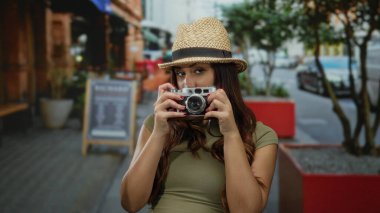 Kameralı genç bir kadın sokakta şapka takıyor. Arka planda ağaçlar ve binalar olan şehir ortamını yakalıyor. Keşif ve macera öneriyor..