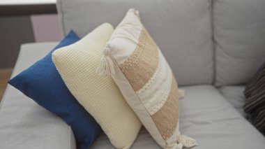 Konforlu oturma odasının içinde çeşitli dekoratif yastıklarla süslenmiş rahat gri bir kanepe var. Mavi, bej ve desenli kumaşlar sergileniyor..