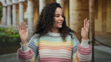 Renkli süveter giyen genç İspanyol kadın, elleri dışarıda, eski bir üniversitede, tarihi mimariyle çevrili, doğal bir şekilde gülümsüyor..