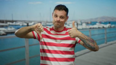 Sahil limanındaki çizgili gömlekli genç adam, açık havada karışık duygular olduğunu düşündüren tekne ve su arkaplanlarına karşı dövmeleri olan baş parmaklarını hem yukarı hem aşağı gösteriyor.