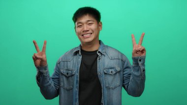 Genç adam rahat kot gömleğiyle gülümsüyor. İki eli de parlak yeşil duvar arkaplanının üzerinde barış işareti yapıyor.