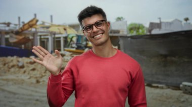 Şehirdeki inşaat alanında kırmızı tişörtlü bir genç açık havada dostça bir jest sergiliyor. Arka planda çekici bir portre sergiliyor..