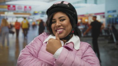 Pembe ceketli, kasklı, havaalanı terminalinde gülümseyen bir kadın güven ve pozitiflik gösteriyor..