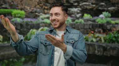 Sakallı genç adam gülümsüyor ve parkta ellerini uzatıyor kot ceket giyiyor açık havada canlı yeşil bir manzara sergiliyor..