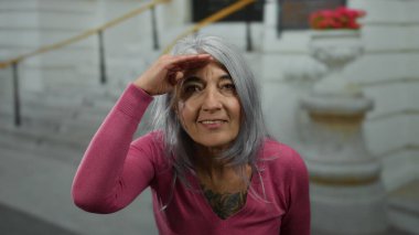 Pembe kazaklı, gri saçlı kadın, eli alnında, arka planda çiçeklerle açık bir sokakta duran mesafeye bakıyor..