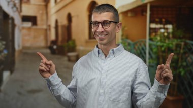 Gülümseyen ve manzaralı sokak ortamında gözlüklü ve çizgili gömlekli bir adam bulanık mimari arka plana karşı işaret ediyor..