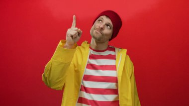 Sarı yağmurluk ve kırmızı bereli genç İspanyol bir adam kırmızı arka plana karşı düşünceli bir hareket yapıyor..