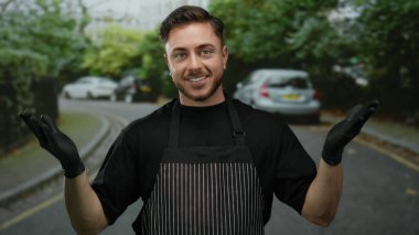 Önlük ve siyah eldiven giyen genç adam park etmiş arabalarla bir caddede gülümseyerek bir garson ya da sokak satıcısı havası yaratıyor..