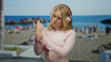 Sarışın, kulaklık takan, deniz kenarında bileğini ovuşturan, sakin bir ifadeyle okyanusun arka planına yaslanmış, rahatlama ve rahatsızlık hissi yakalayan bir kadın..