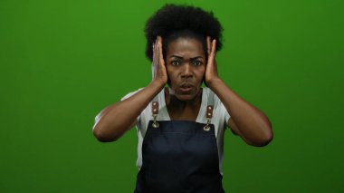 Önlüklü kadın, elleri başının üstünde yeşil arka plana karşı duruyor. Stres ve şaşkınlık taşıyor, beyaz gömlek ve küpeler takıyor..