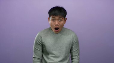 Şaşırmış bir ifadeye sahip genç bir adam, izole bir mor arka plana karşı duruyor, gri bir gömlek giyiyor, bir anlık şok ve duyguyu özetliyor..