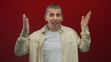 Şaşırmış ifadesi olan bir adam, sıradan bir bej tişört ve beyaz tişört giymiş, kırmızı bir duvarın önünde gözleri ortaya çıkarıyor. Beklenmedik duygular taşıyor..