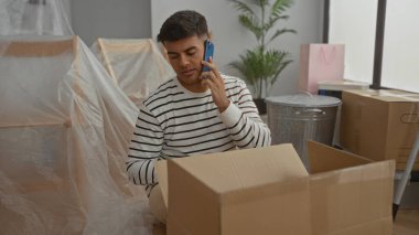 Çizgili gömlekli genç adam, yeni bir apartman dairesinde karton kutuları açarken telefonla konuşuyor. Yeni bir taşınma ya da taşınma işlemi öneriyor..