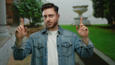 Sakallı genç adam şehir dışında bir sokak ortamında elini kaldırıyor, kot ceket giyiyor, düşünceli bir bakışla jest yapıyor, yemyeşil ve şehir mimarisi ile çevrili..