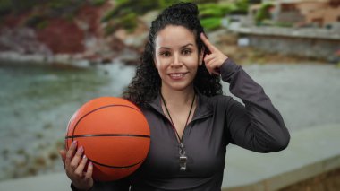 Plaj sahilinde basketbol oynayan bir kadın, boynunda ıslık çalan, açık gökyüzünün altında rahat ve düşünceli bir ifade sergiliyor..