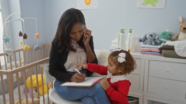 Bebek beşiğinin yanında panoya yazı yazan küçük kızı olan bir kadın yatak odasında oyuncaklarla ve temel ihtiyaçlarla çevrili.