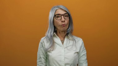 Gri saçlı ve gözlüklü yaşlı bir kadın turuncu bir arka plan duvarına karşı neşeli bir yüz çiziyor, beyaz bir gömlek giyiyor ve garip bir ifadeyle yukarı bakıyor..
