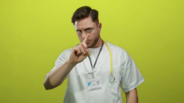 Sakallı, beyaz, genç bir doktor. Parmak ucunda, izole edilmiş sarı arka plana karşı steteskop takıyor. Ciddi bir ifade ve profesyonel bir varlık sergiliyor..