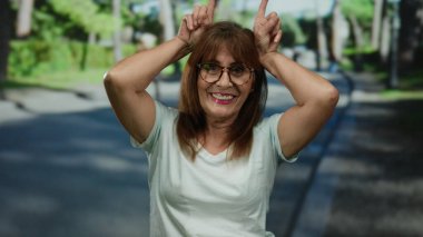 Kıdemli İspanyol kadın neşeli bir gülümseme ve gözlüklerle güneşli bir günde neşe içinde parkta poz veriyor..
