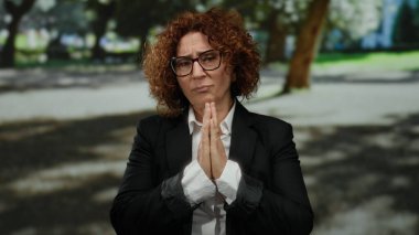 Kıvırcık saçlı gözlüklü bir kadın güneşli bir parkta dua ediyor. Huzurlu bir ortam sergiliyor..