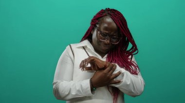 Örgülü ve gözlüklü bir kadın bileğini izole edilmiş yeşil bir duvar arkasına yaslar ve bilek bölgesindeki acıyı ifade eder..