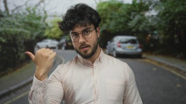 Sakallı genç adam, ağaçlarla çevrili bir sokakta jest yapıyor. Ve gözlüklü ve açık renk gömlekli park edilmiş arabalar.