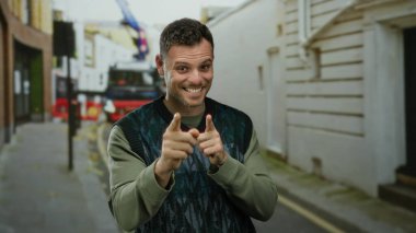 Sakallı genç bir adam, kenar mahallede kendine güvenerek gülümsüyor. Açık havada moda anlayışı sergiliyor. Olumlu hareketler yapıyor..