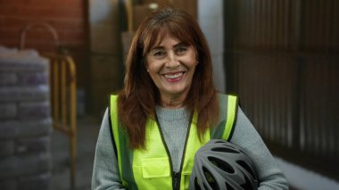 İçeride inşaat alanında gülümseyen, güvenlik yeleği giyen ve arka planda endüstriyel malzeme ve aletlere karşı kask tutan İspanyol kıdemli kadın..