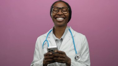 Doktor gülümserken pembe arka planda cep telefonu kullanıyor, steteskop takıyor, büyük gözlük takıyor, beyaz önlük takıyor, profesyonel iletişim ve mutluluk taşıyor.