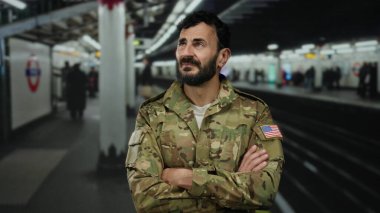 Askeri üniformalı bir adam, bir yeraltı istasyonunda kollarını kavuşturmuş, bulanık banliyö ve metro işaretleri arasında düşünceli bir yüz ifadesi sergiliyor, kentsel geçiş modunu vurguluyor..