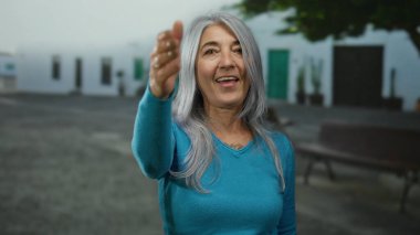 Gri saçlı yaşlı bir kadın, sokakta dururken elini uzatıyor, açık havada gülümsüyor, açık hava kentsel ortamında dostça bir sokak etkileşimi sergiliyor..