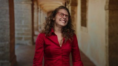 Kırmızı tişörtlü, kıvırcık saçlı ve gözlüklü bir kadın eski üniversite koridorunda içeride dikiliyor..