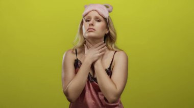 Pembe gecelik elbiseli sarışın kadın sarı arka plandan rahatsız görünüyor, birden fazla ifade ile boyuna dokunuyor, rahatsızlık ve duygu taşıyor..